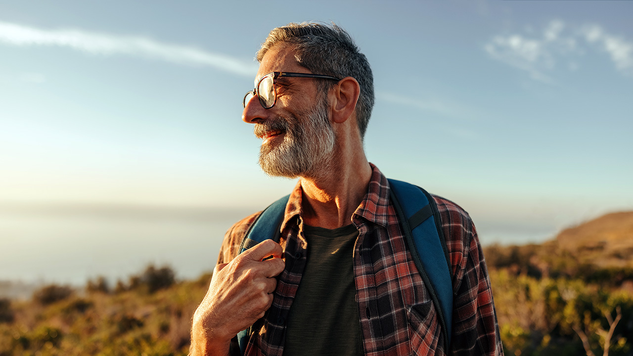 A bearded man with glasses gazes into the distant landscape, wearing a blue backpack