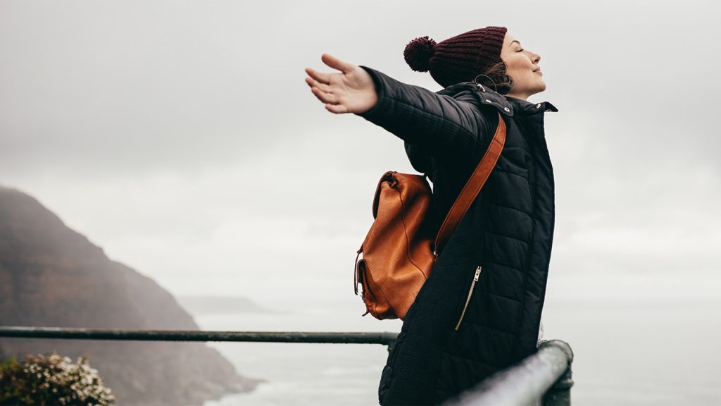 A woman wearing a puffer jacket, orange backpack and beanie leans forward over a scenic view, arms outstretched